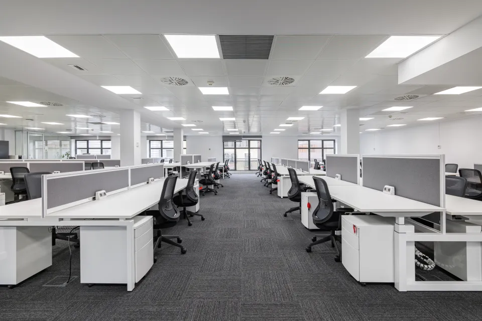 Large empty open-plan office with rows of white desks and black chairs under bright fluorescent lighting